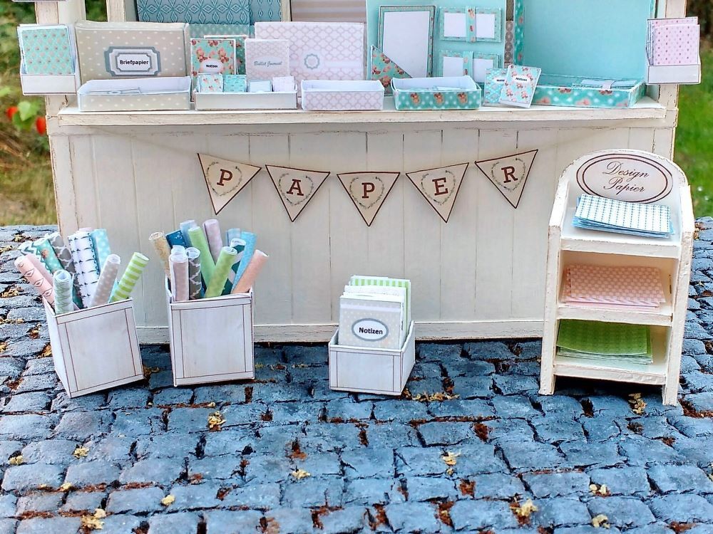Miniatur-Bastelstand mit pastellfarbenen Designpapieren, Papierrollen und Notizblöcken