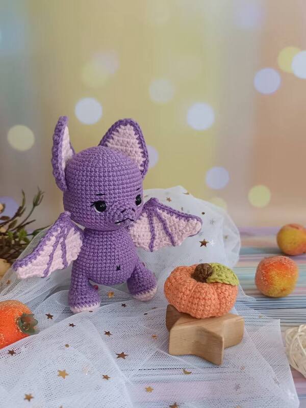 Purple crocheted amigurumi bat with pink-lined wings next to a small crocheted pumpkin on tulle with star confetti