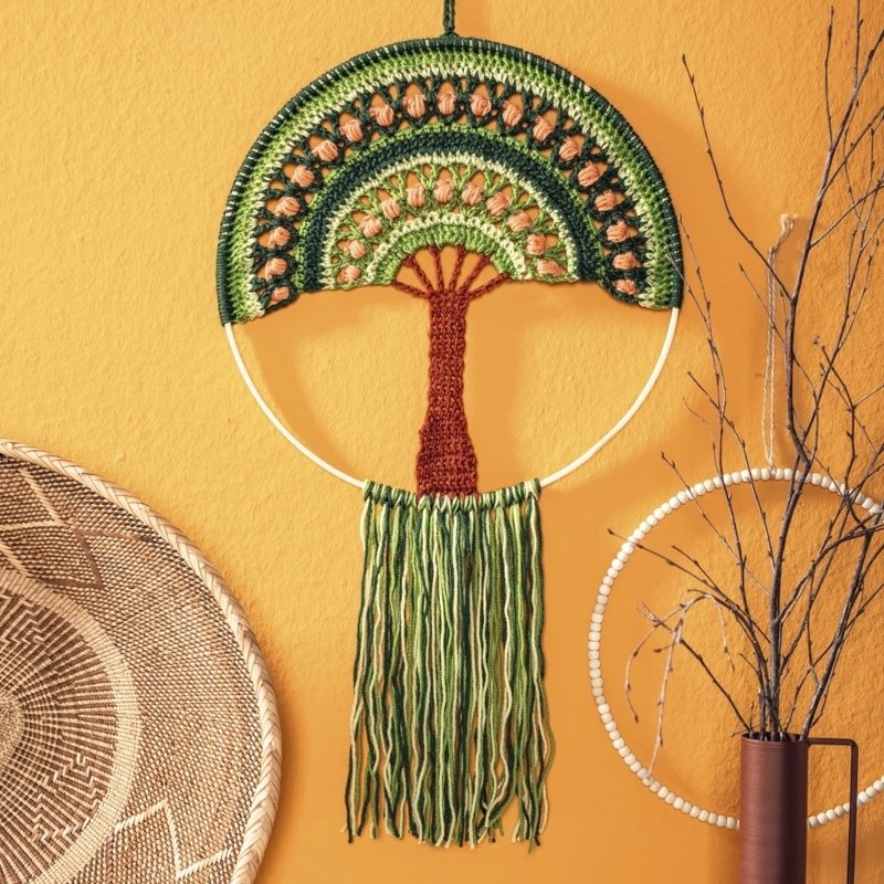 Gehäkelter Wandbehang auf Ring in Lebensbaum-Form mit grüner Krone, braunem Stamm und grünen Fransen vor warmgelber Wand