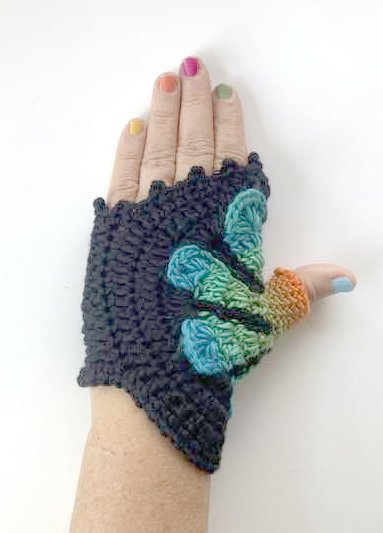 Hand mit dunkelgrauer gehäkelter fingerloser Stulpe und blau-grüner Schmetterlings-Applikation, bunte Fingernägel