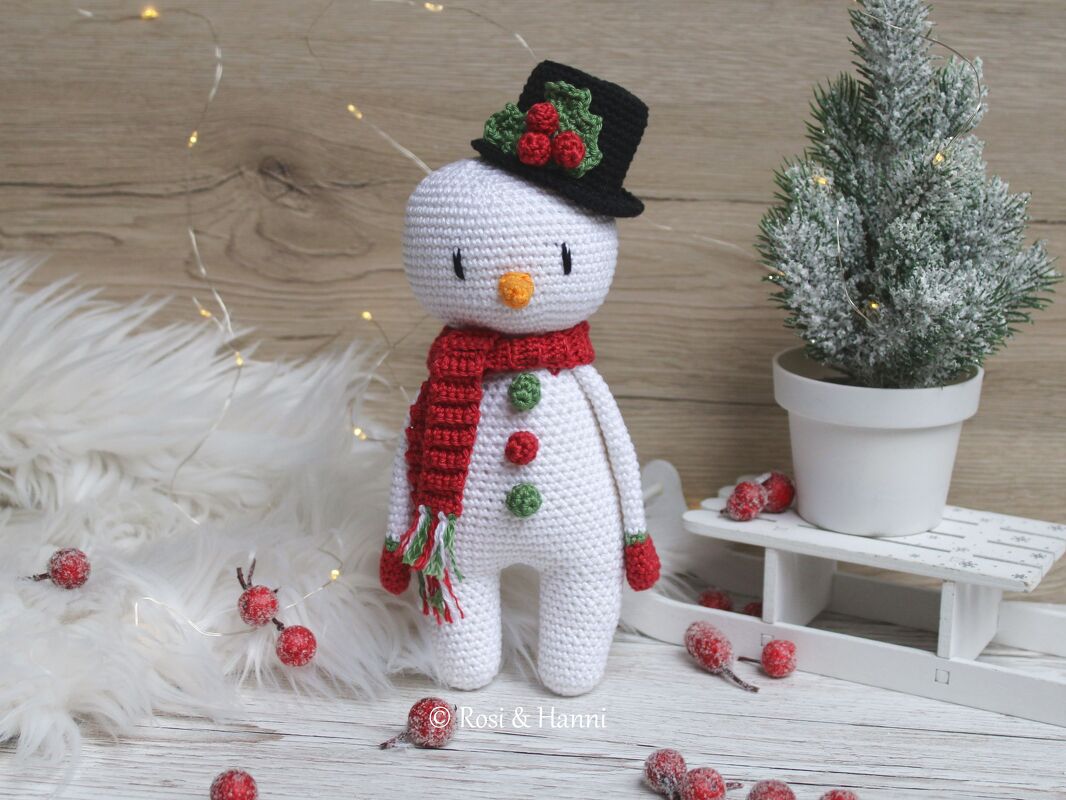 Gehäkelter Schneemann-Amigurumi mit schwarzem Zylinder, rotem Schal und bunten Knöpfen, umgeben von winterlicher Dekoration