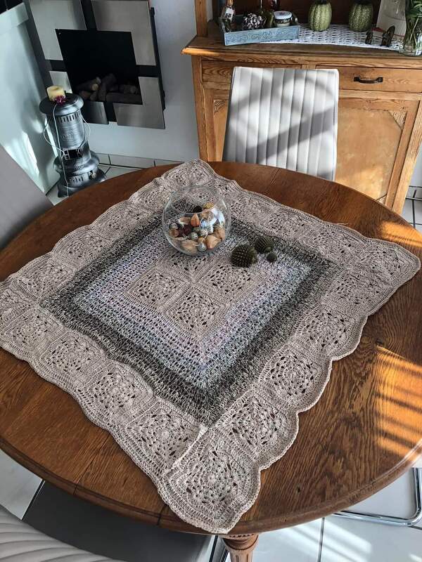Quadratisches gehäkeltes Deckchen in Beige- und Grautönen auf einem Holztisch, dekoriert mit einer Glasschale mit Muscheln und kleinen Dekokakteen.