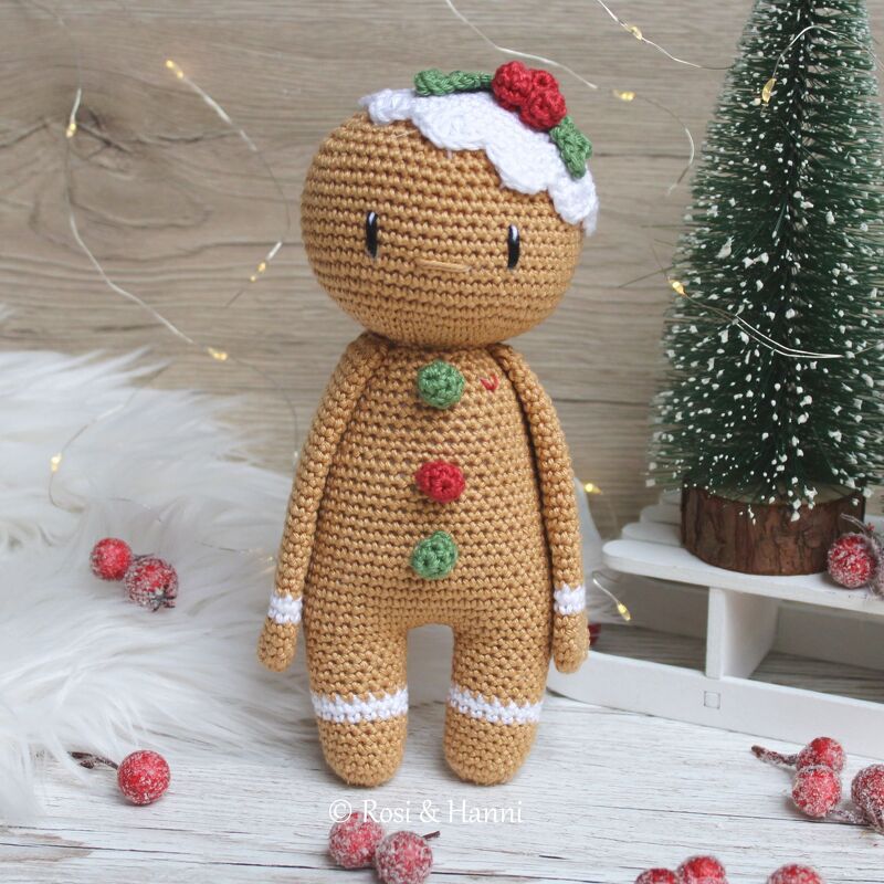 Gehäkelter Lebkuchenmann mit Zuckerguss, roten Beeren und grünen Knöpfen neben Mini-Weihnachtsbaum