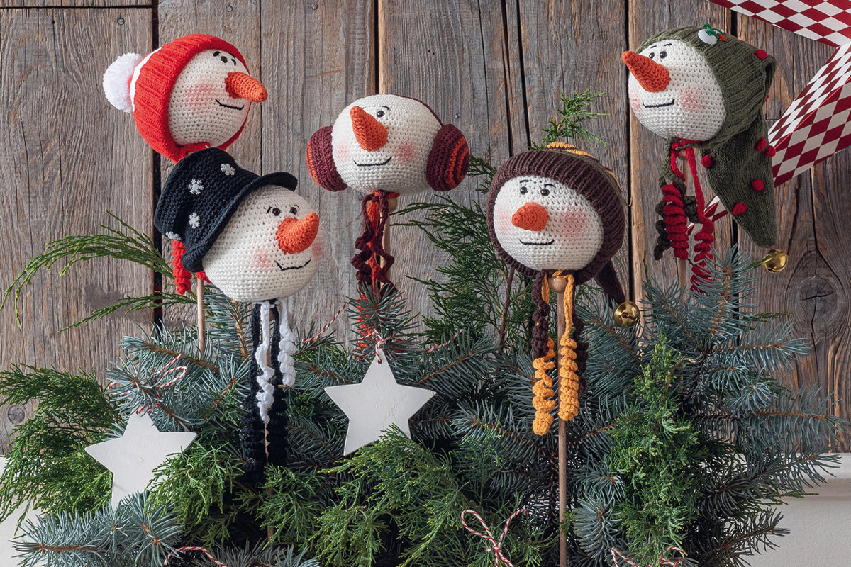 Fünf gehäkelte Schneemannköpfe mit Mützen auf Holzstäben im Tannengrün vor Holzplanken