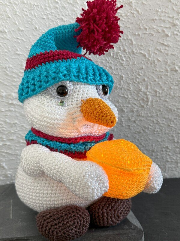 Gehäkelter Schneemann mit türkis‑roter Mütze, Schal und orangefarbener Leuchte
