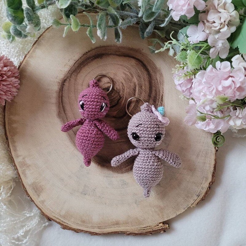 Zwei gehäkelte Amigurumi-Schlüsselanhänger in Rosé und Flieder auf einer Holzscheibe