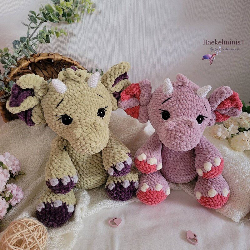 Zwei sitzende gehäkelte Amigurumi‑Drachen in Pastell (beige‑violett und rosa‑lila) mit Hörnern und kontrastierenden Pfoten