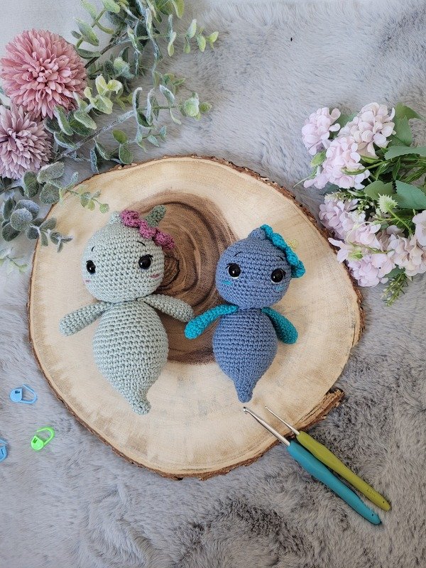Zwei Amigurumi-Figuren in Mintgrün und Blau auf einer Holzscheibe, daneben Häkelnadeln und Maschenmarkierer, umgeben von Blüten.