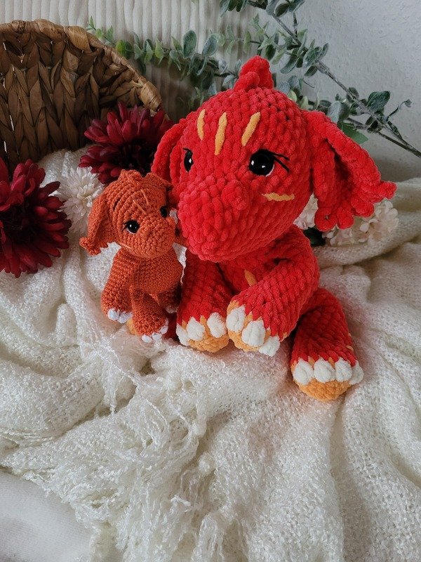 Red chenille-style crochet dragon with cream claws and yellow markings beside a small orange amigurumi on a cream knit blanket