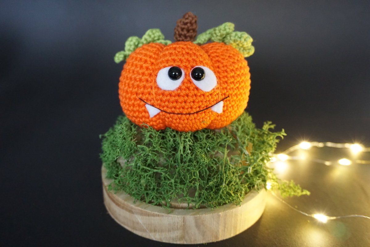 Gehäkelter orangefarbener Amigurumi-Kürbis mit Gesicht auf Moos und Holz, beleuchtet von Lichterketten