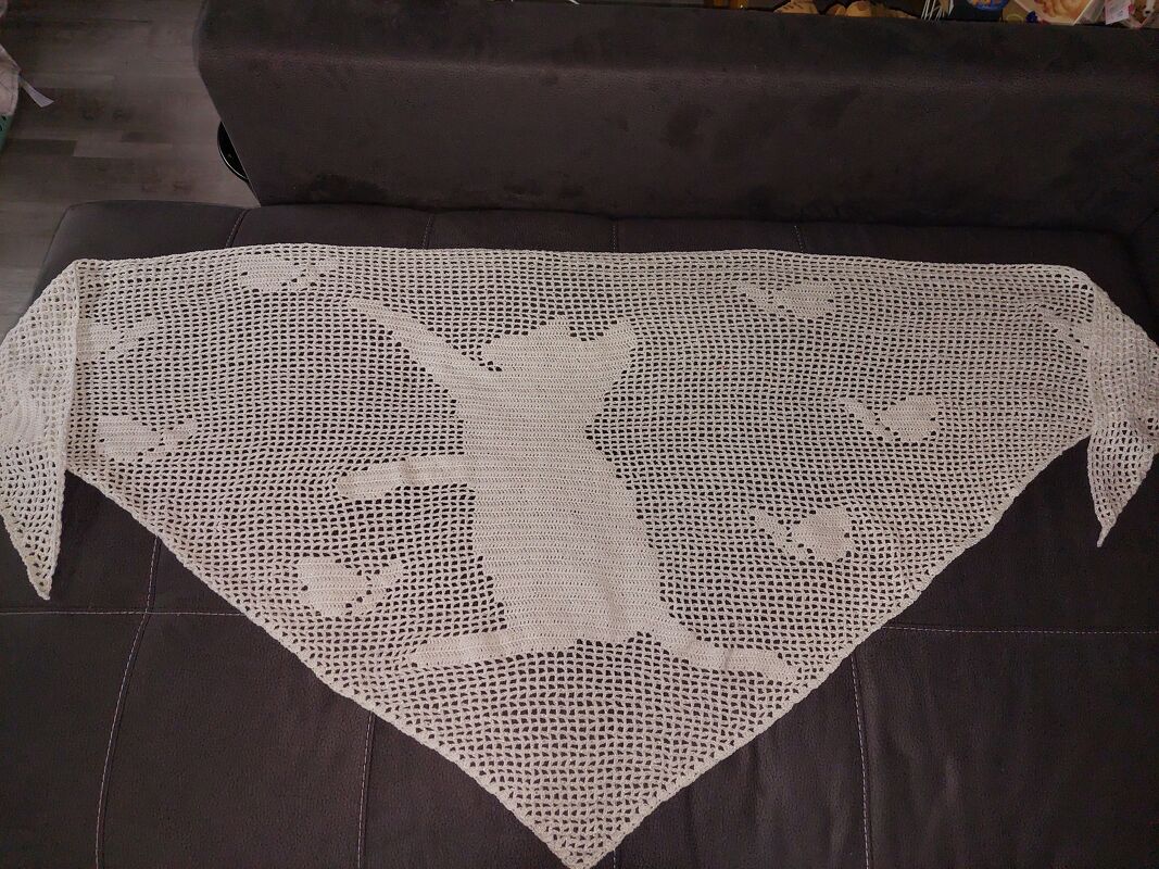 Helles, handgehäkeltes Dreieckstuch in Filethäkeltechnik mit großer Katzen-Silhouette und kleinen Vogelmotiven, ausgebreitet auf dunklem Sofa.