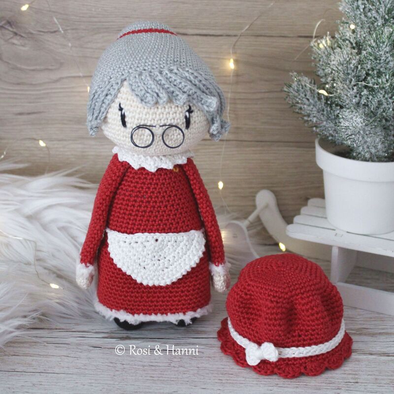 Gehäkelte Weihnachts-Oma Amigurumi in rotem Kleid mit weißer Schürze, grauem Haardutt und rotem Hut neben der Puppe