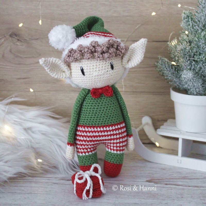 Gehäkelter Weihnachtself in Grün und Rot mit Zipfelmütze, roter Fliege und gestreifter Hose neben kleinem roten Geschenk vor weihnachtlicher Dekoration.