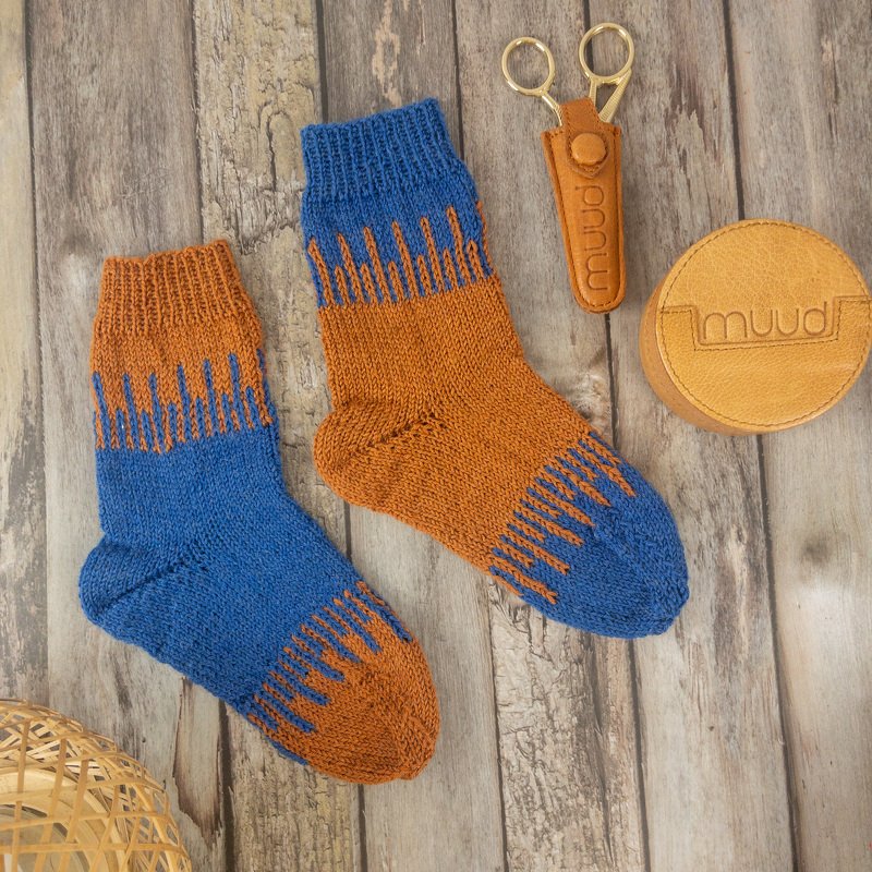Zwei handgestrickte Socken in Blau und Rostorange mit Zackenmuster auf Holzuntergrund.