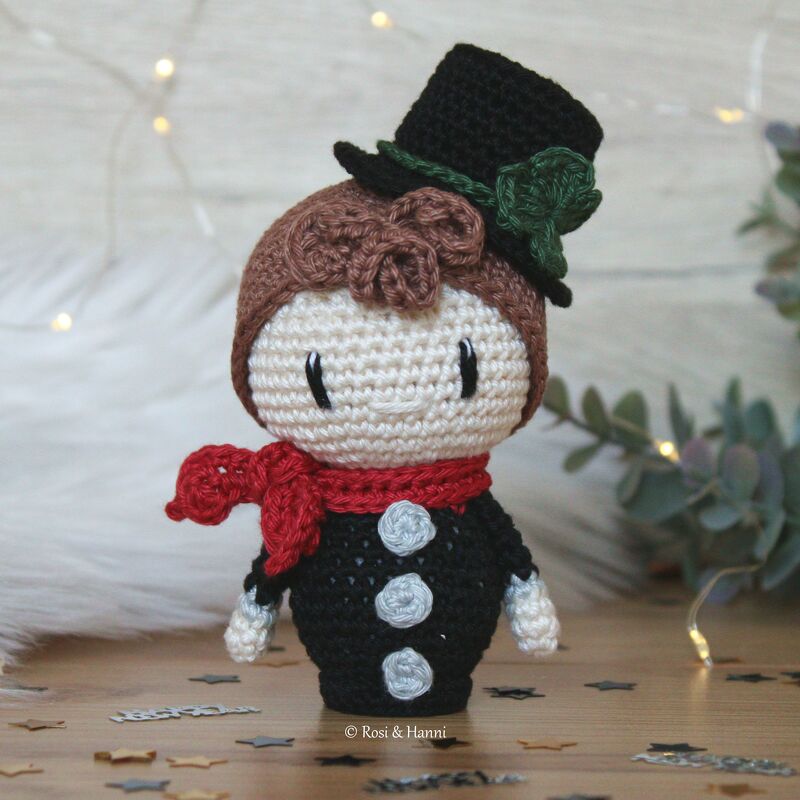 Gehäkelte Amigurumi-Puppe mit braunem Haar, schwarzem Zylinder mit grünem Band, rotem Schal und schwarzem Mantel mit drei hellgrauen Knöpfen auf einem Holztisch mit Sternkonfetti und Lichterketten.