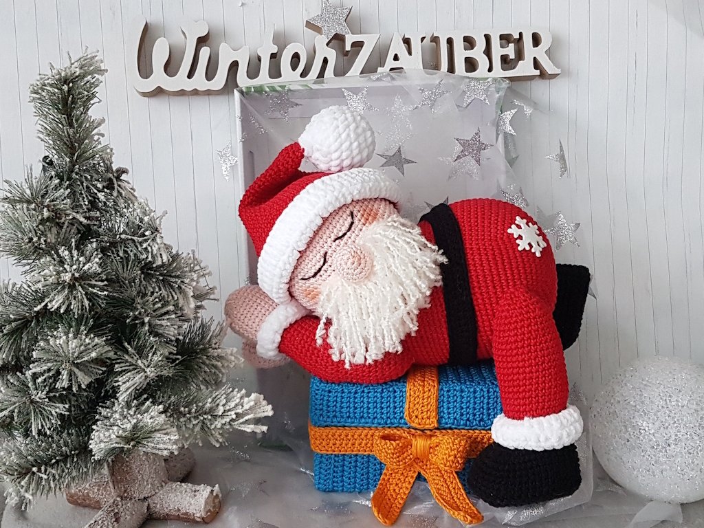 Gehäkelter Weihnachtsmann in roter Mütze schläft auf blauem Geschenk neben beschneitem Tannenbaum