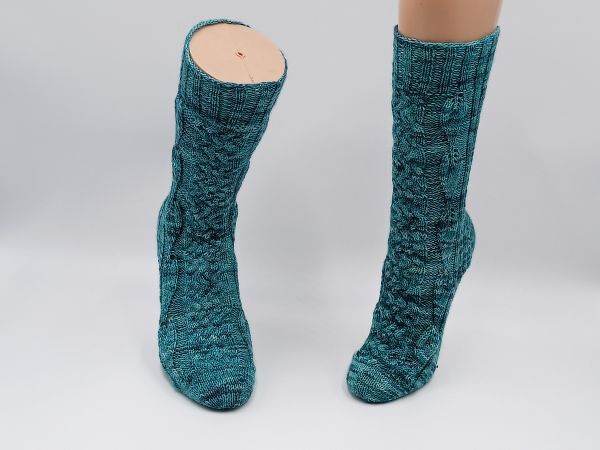 Zwei gestrickte türkise Zopfsocken mit Zopf- und Rippenmuster, eine an einem Bein präsentiert.