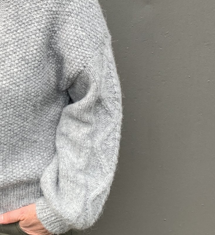 Detailaufnahme eines hellgrauen Damenpullovers mit Perlmuster am Rumpf und Zopfdetail am Ärmel