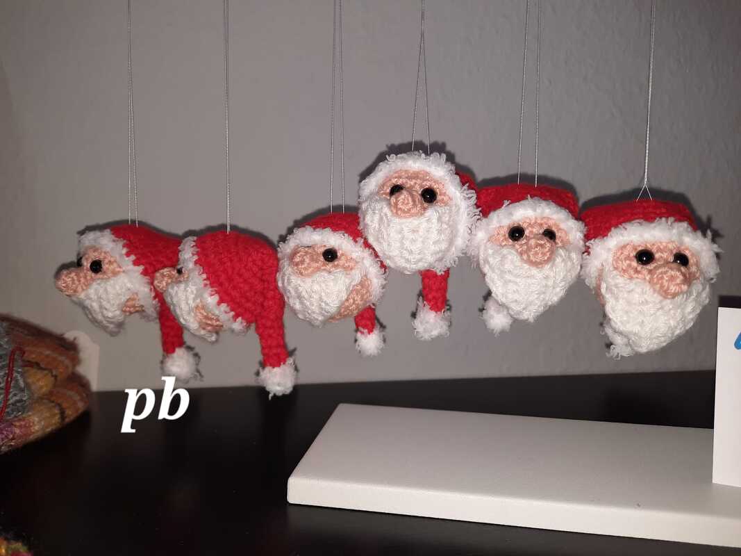 Fünf kleine gehäkelte Weihnachtsmann-Köpfe mit roten Mützen und weißen Bärten an Fäden hängend.