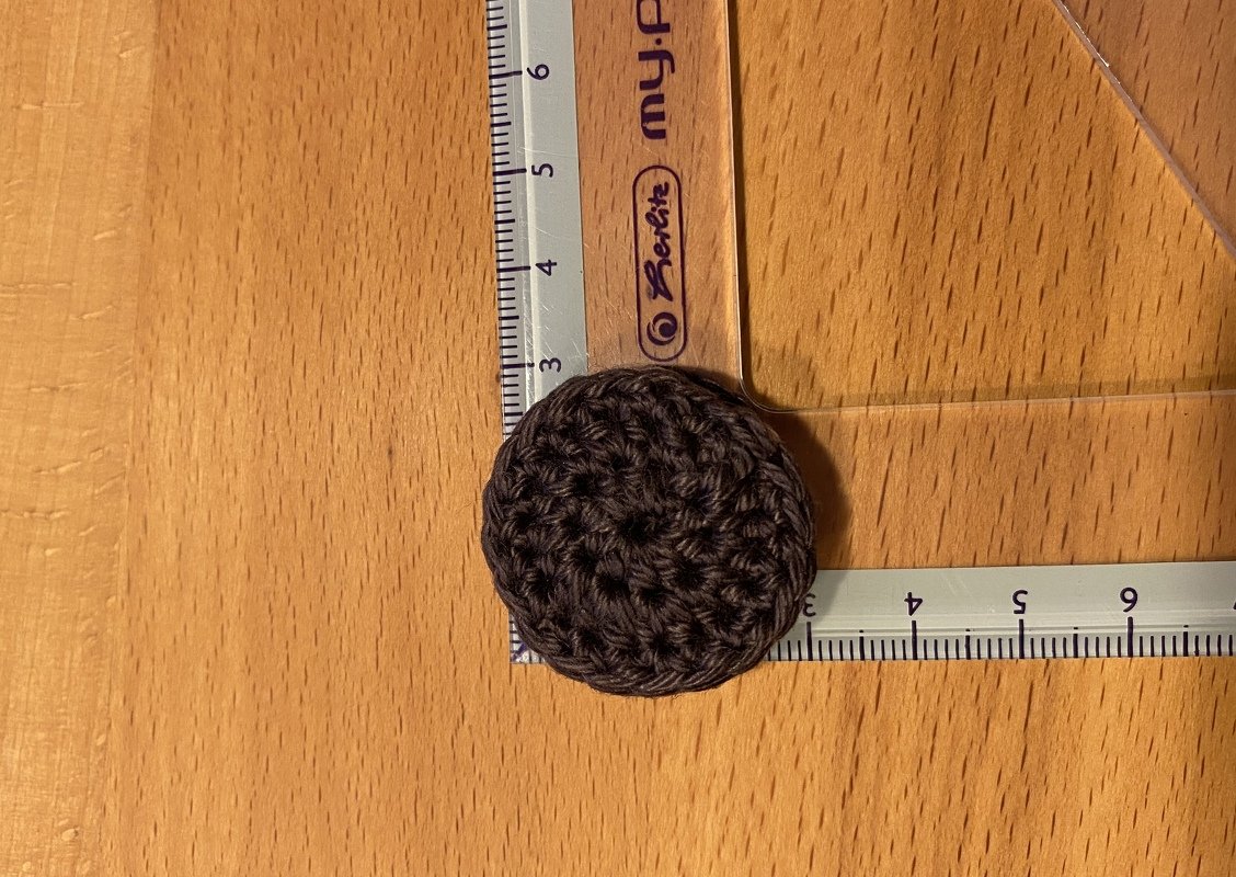 Kleines rundes dunkelbraunes Häkelstück neben einem Lineal, etwa 3 cm Durchmesser