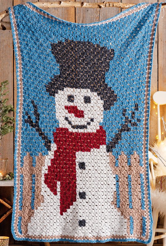Blaue gehäkelte Decke mit großem weißen Schneemann, schwarzem Zylinder, rotem Schal, braunen Armen und kleinem braunen Zaun am unteren Rand.