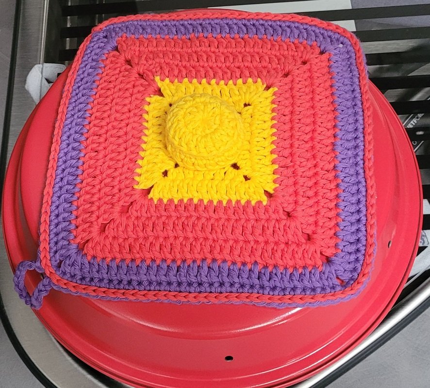 Quadratischer gehäkelter Topflappen mit gelbem Mittelkreis, korallroten Reihen, lila Rand und Aufhängeöse