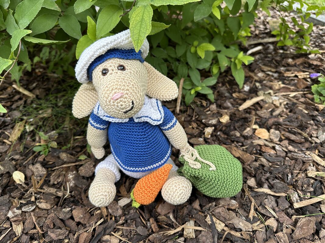 Gehäkeltes Amigurumi-Schaf im Matrosenlook mit Mütze, Karotte und grünem Beutel vor grünen Blättern