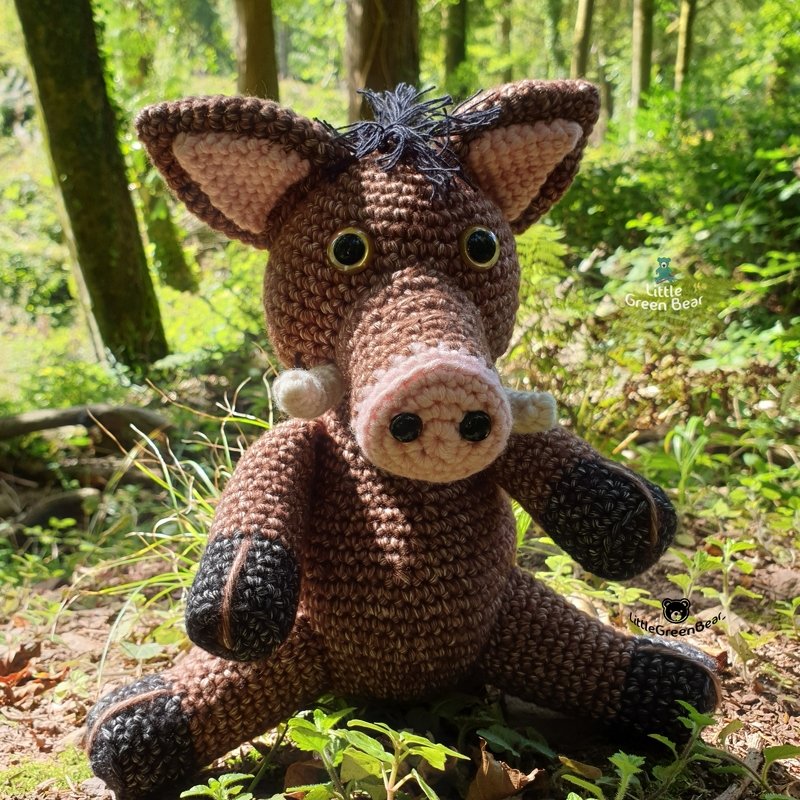 Crochet amigurumi boar with long snout and tusks sitting on the forest floor