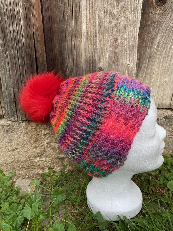 Bunte gehäkelte Mütze mit rotem Bommel auf einem Styroporkopf vor Holz und Gras