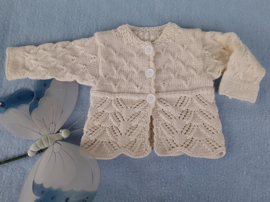 Cremefarbene gestrickte Babyjacke mit drei weißen Knöpfen, Zopf- und Ajourmuster auf blauem Untergrund