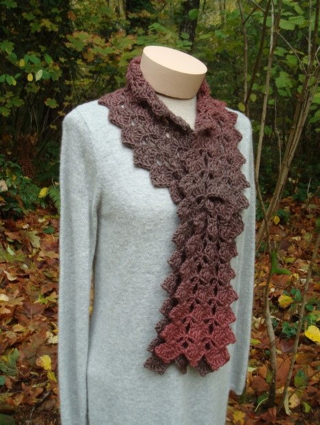 Brown crocheted scarf with leaf-like scalloped pattern on a mannequin in an autumn woodland setting.
