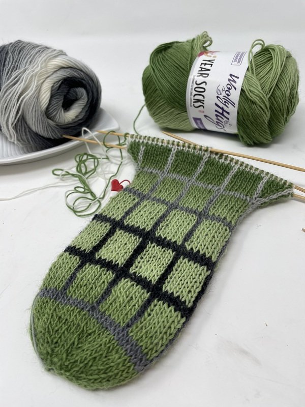 Halbfertige grüne Socke mit schwarz-grauem Karomuster und Garnknäuel