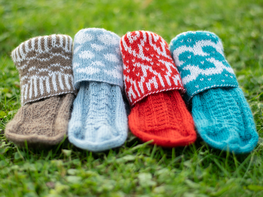 Four folded knitted socks in brown, light blue, red and teal laid side by side on grass, showing various stitch patterns and patterned cuffs