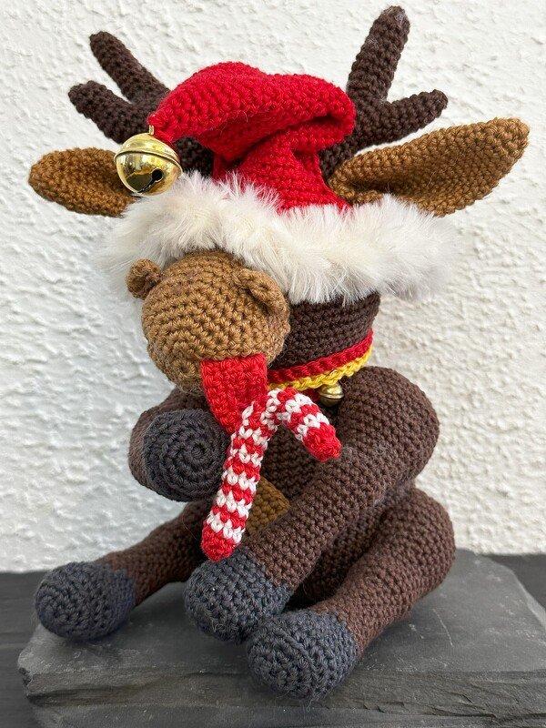Gehäkeltes braunes Rentier-Amigurumi, sitzend, mit roter Mütze, weißem Fellrand, Glöckchen und rot-weiß gestreiftem Schal