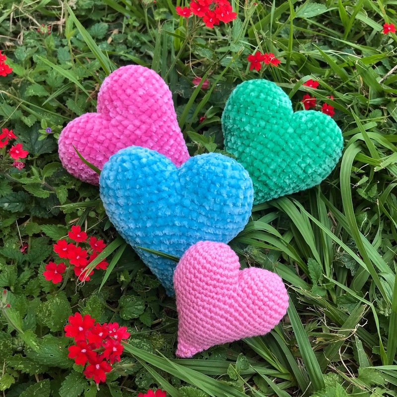 Vier Amigurumi-Herzen in verschiedenen Größen und Farben auf einer Wiese mit roten Blumen