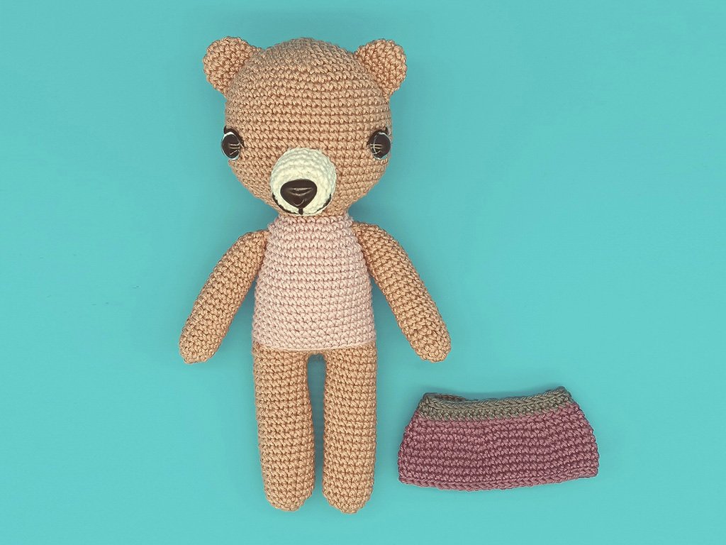 Gehäkelter Amigurumi-Teddy in Beige mit hellrosa Oberteil; daneben ein gehäkelter Rock in Altrosa auf türkisfarbenem Hintergrund