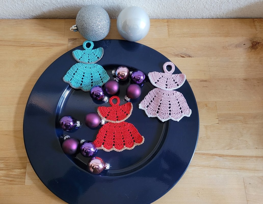 Drei gehäkelte Engelkleider in Türkis, Rosa und Rot auf dunklem Teller mit violetten und silbernen Weihnachtskugeln