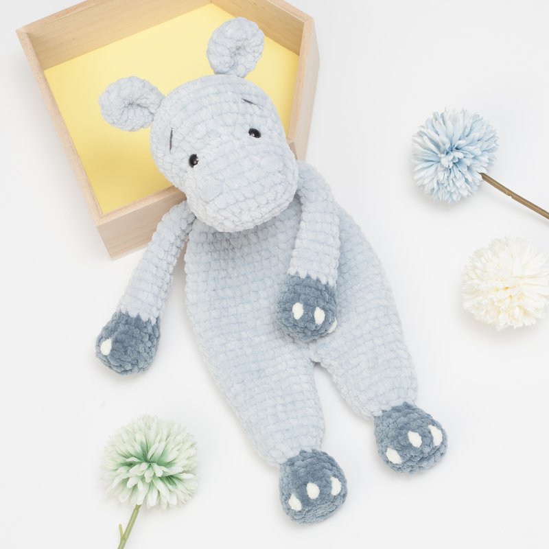 Light blue crocheted hippo snuggle blanket with darker blue spotted paws, shown on a white background with a wooden box and decorative flowers.