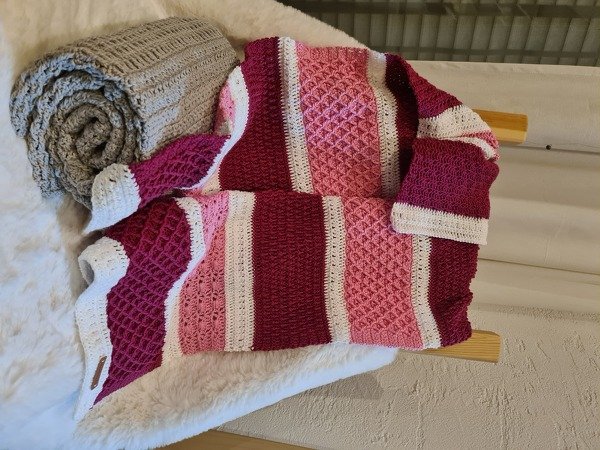 Handgehäkelte Babydecke mit Streifen in Rosa, Weiß und Bordeaux, dazu gerollte graue Decke.