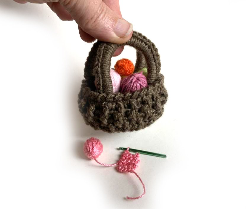 Hand hält einen kleinen braunen Häkelkorb mit bunten Garnknäueln; daneben Häkelnadel und angefangene rosa Häkelarbeit.