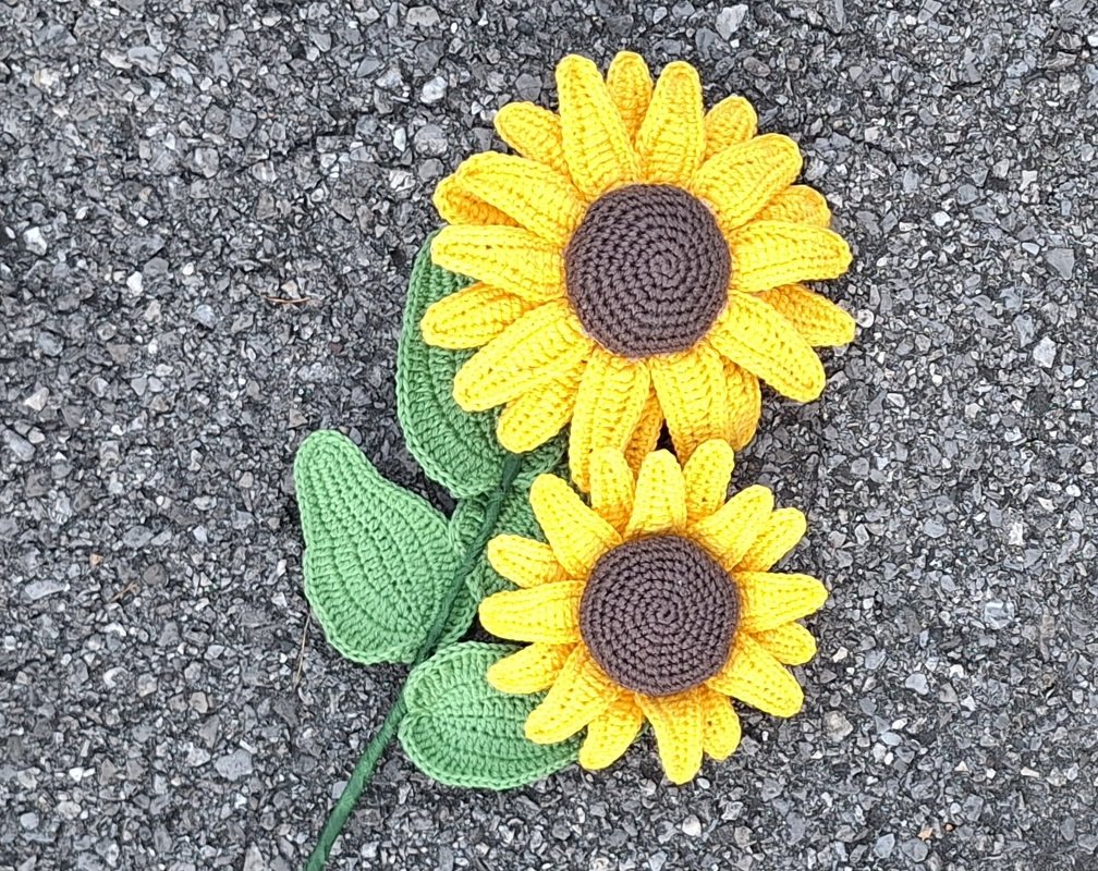 Zwei gehäkelte gelbe Sonnenblumen mit braunen Mittelteilen und grünen Blättern auf grauem Kiesgrund.