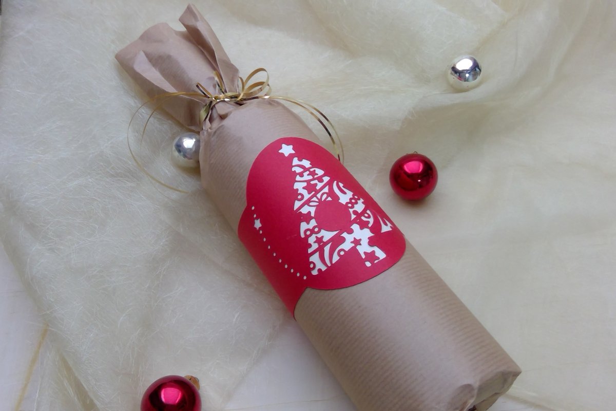 Flasche in braunem Packpapier mit roter Banderole und ausgeschnittenem Tannenbaum‑Motiv, goldene Schleife und Weihnachtskugeln