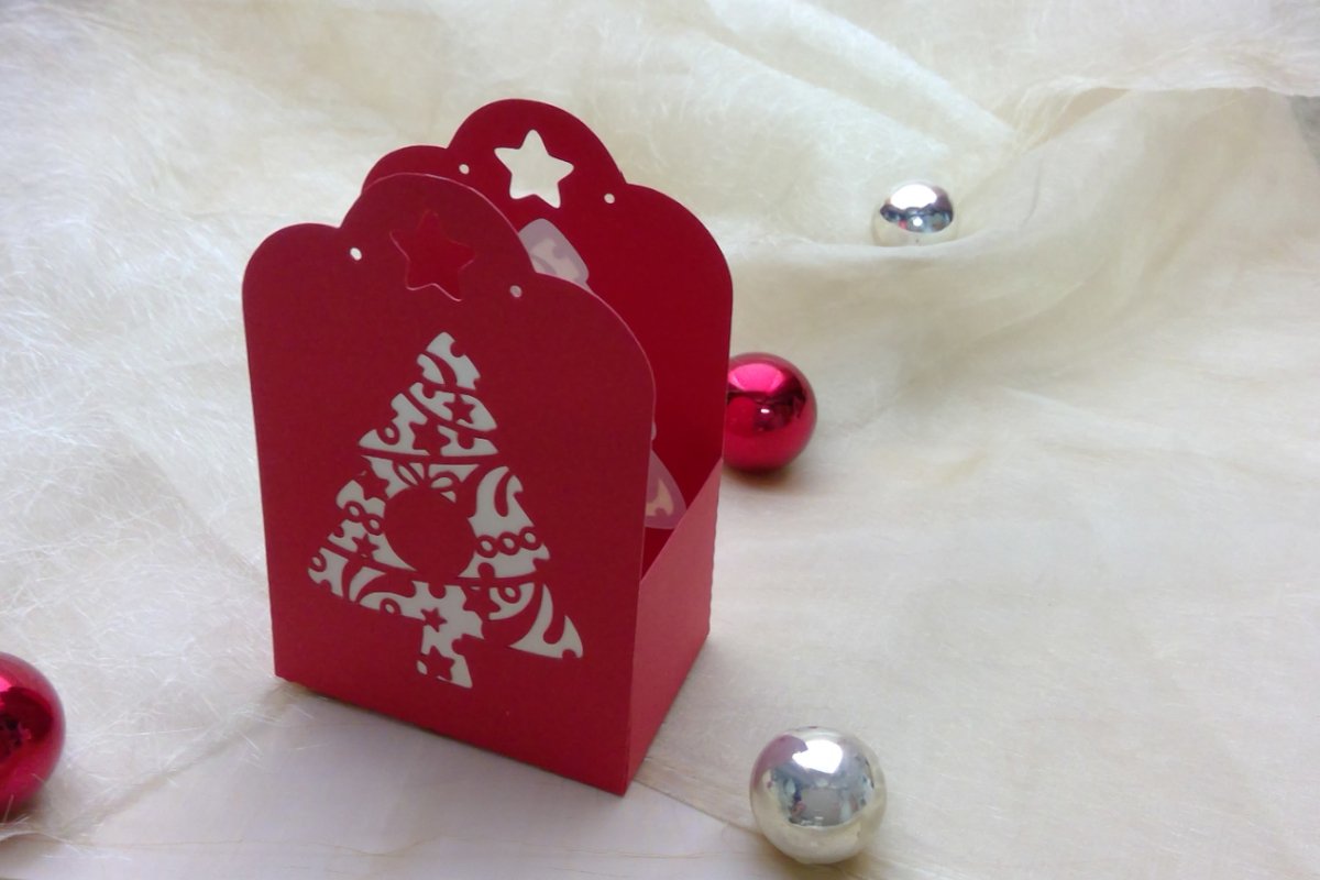 Rote Geschenkbox mit ausgeschnittenem Tannenbaum und Sternen, umgeben von roten und silbernen Kugeln auf hellem Stoff