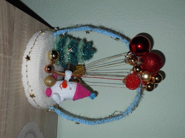 Gehäkelter Schneemann mit rosa Mütze vor kleiner Tanne auf rundem Sockel, umgeben von roten Kugeln und blauem Glitzerbogen