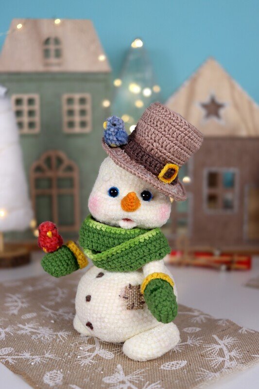 Crocheted snowman amigurumi with a brown top hat, blue and red birds, green scarf and mittens.