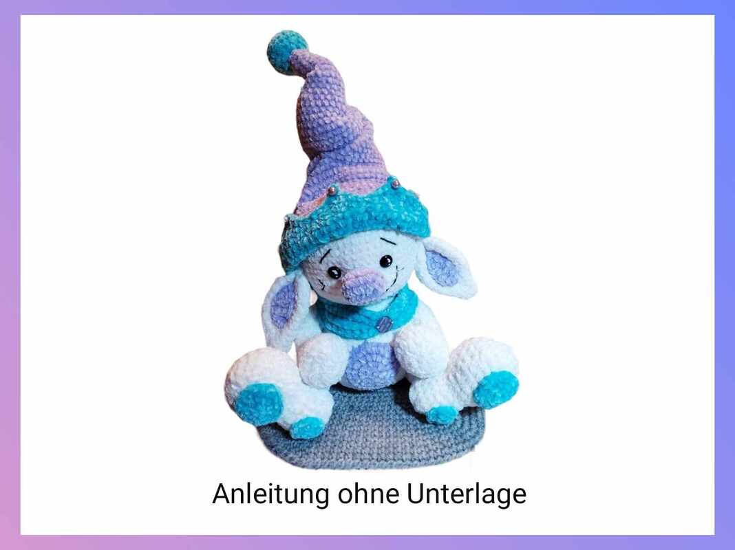 Weißer gehäkelter Amigurumi-Hase mit lila- und türkisfarbener Zipfelmütze und Schal auf grauem Untergrund.