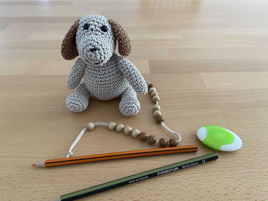 Kleiner gehäkelter Hund mit braunen Schlappohren neben zwei Buntstiften, Radiergummi und Holzperlen auf Holztisch.
