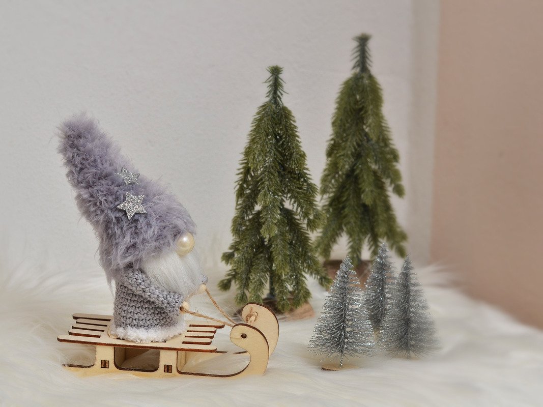 Gehäkelter Wichtel mit flauschiger Sternmütze auf Holzschlitten neben Mini-Tannen
