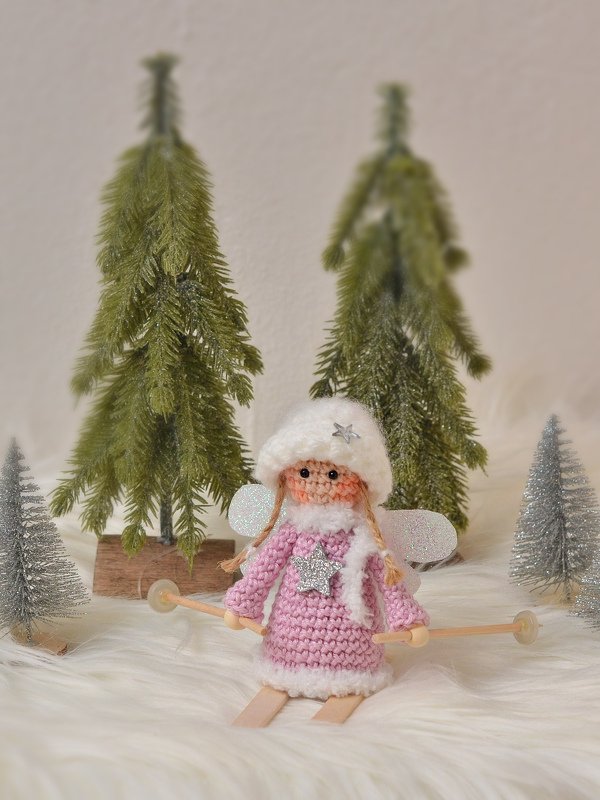 Gehäkeltes Amigurumi-Engelchen in rosa Kleid und weißer Mütze mit Mini-Skiern vor Deko-Tannen