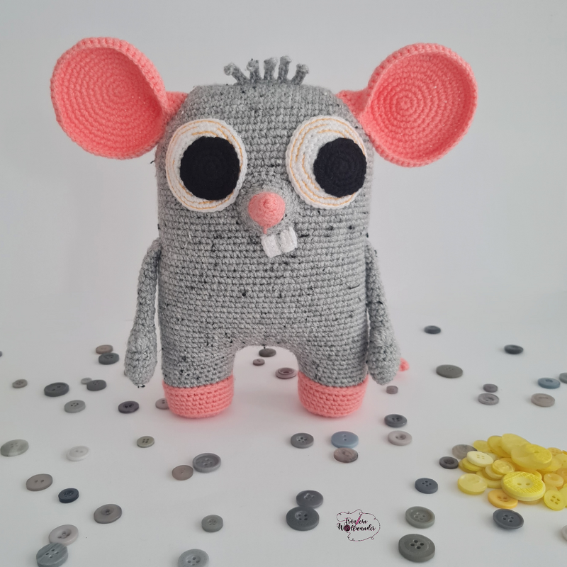 Gehäkelte graue Amigurumi-Maus mit rosa Ohren und Pfoten, großen schwarzen Augen und zwei weißen Zähnen, davor verstreute Knöpfe